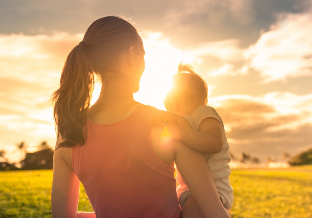 View from behind of a young woman holding a small child looking into a bright, golden sunset